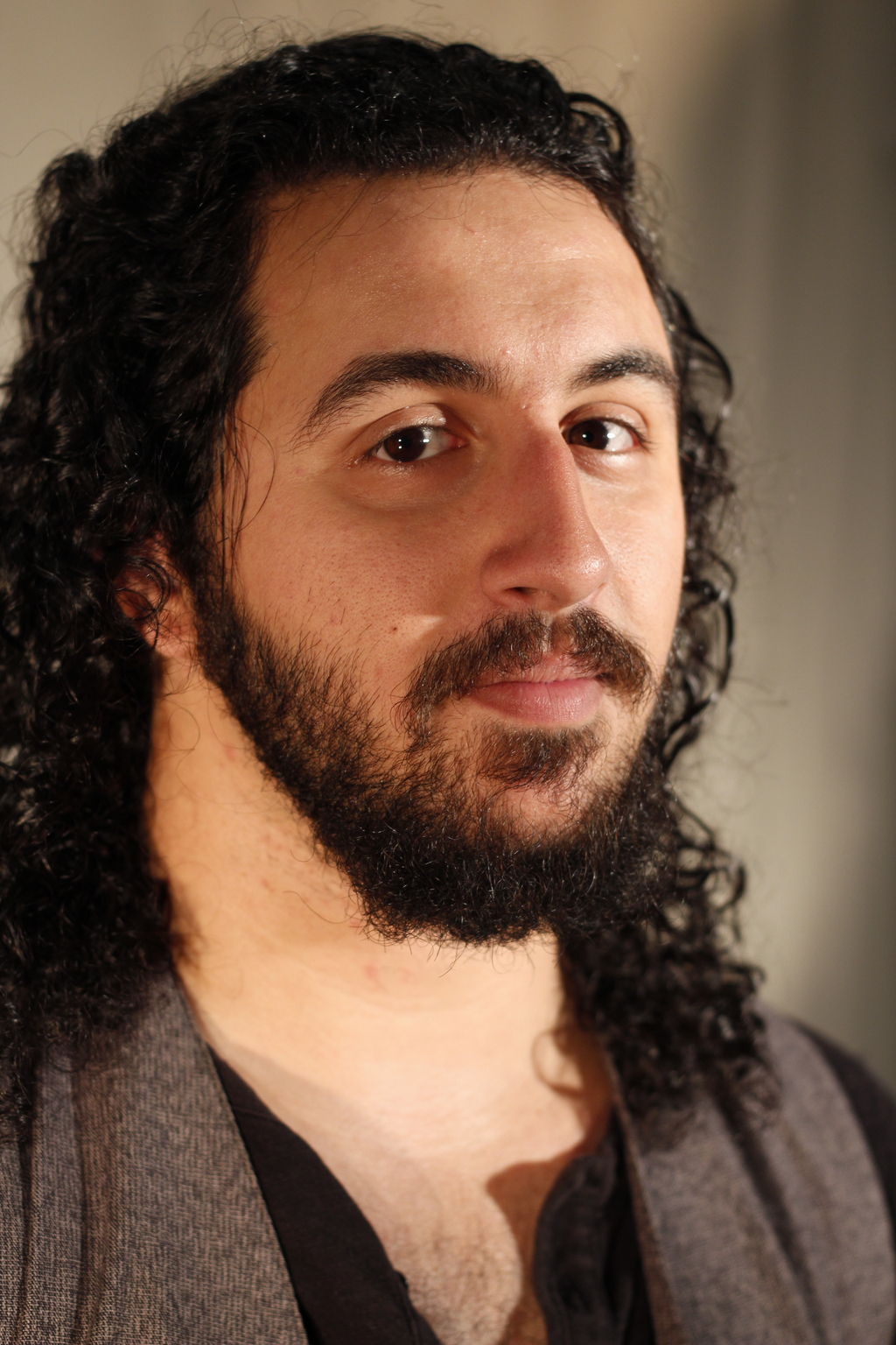 an actor with long black hair and a black beard wearing a black shirt and grey vest looks at the camera in 3/4 profile